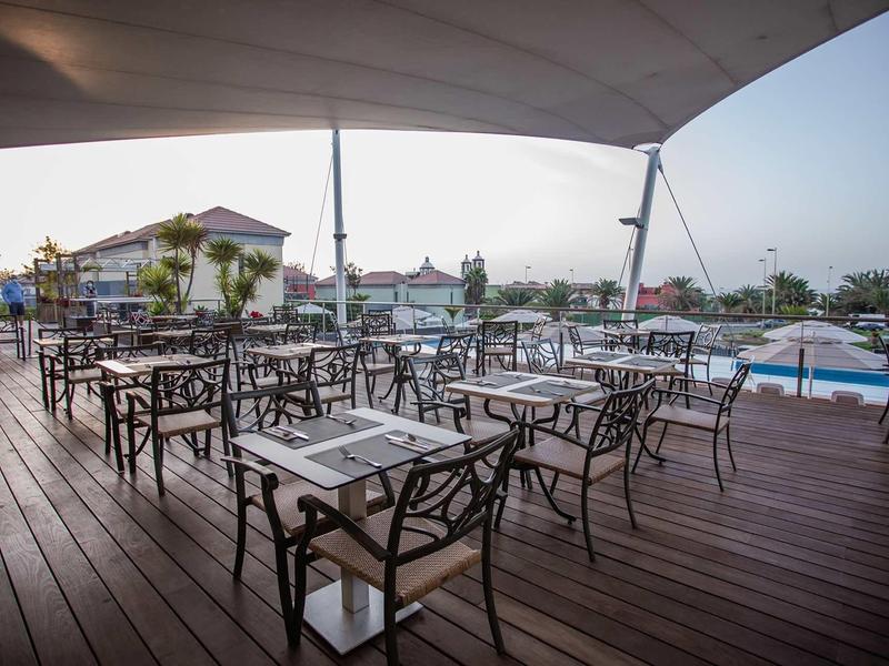 Offene Außenterrasse mit Holzboden, Metallstühlen und Tischen unter großem Sonnensegel.