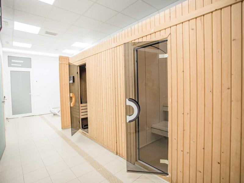 Heller Raum mit Holzwand und Glastür, die Zugang zu einer Sauna bietet. Weißer Fliesenboden.