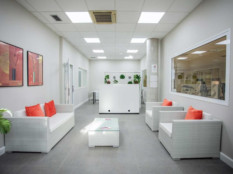 Heller Wartebereich mit weißen Sofas, orangen Kissen, Pflanzen und großen Fenstern zur Office-Seite.