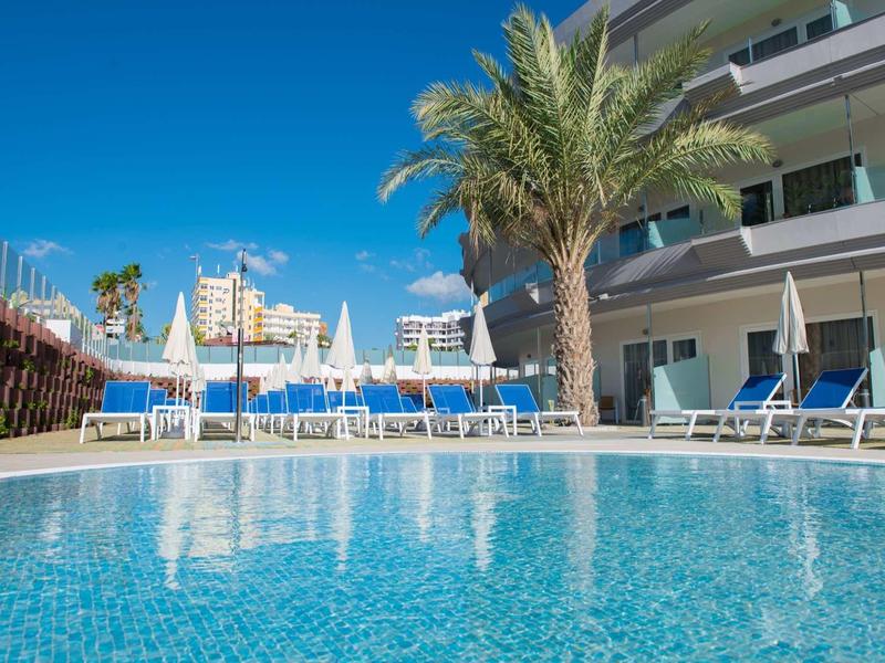 Piscine bleue claire avec transats et palmiers à côté du bâtiment de l'hôtel sous un ciel bleu.