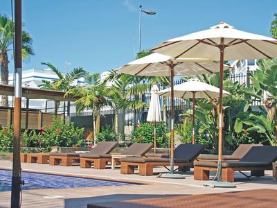 Poolbereich mit Sonnenliegen, großen Sonnenschirmen und tropischer Bepflanzung bei klarem Himmel.