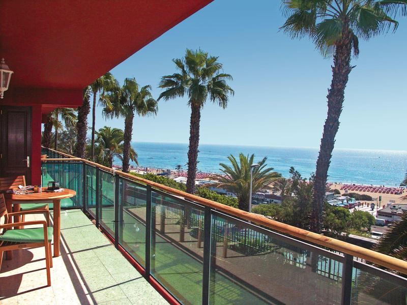 Balkon mit Glasgeländer, Holzmöbeln und Palmenblick auf das blaue Meer bei klarem Himmel.