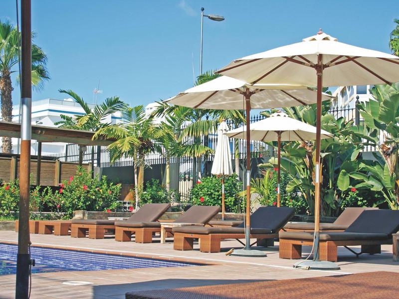 Poolbereich mit Sonnenliegen, großen Sonnenschirmen und tropischer Bepflanzung bei klarem Himmel.