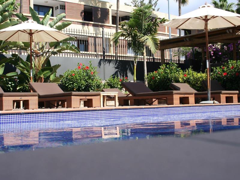 Blick auf einen blauen Pool mit braunen Sonnenliegen und Sonnenschirmen vor einer weißen Wohnanlage.