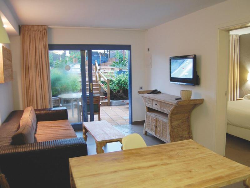 Gemütliches Wohnzimmer mit Couch, Holztisch, TV, Balkon und Blick auf Garten und Pool.