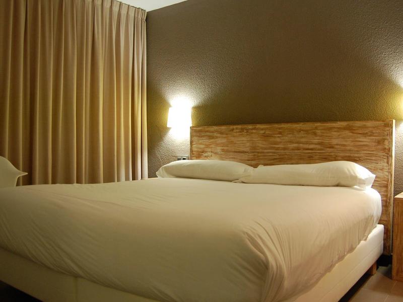 Ein schlichtes Schlafzimmer mit großem Bett, Holzkopfteil, zwei weißen Kissen und warmem Licht.