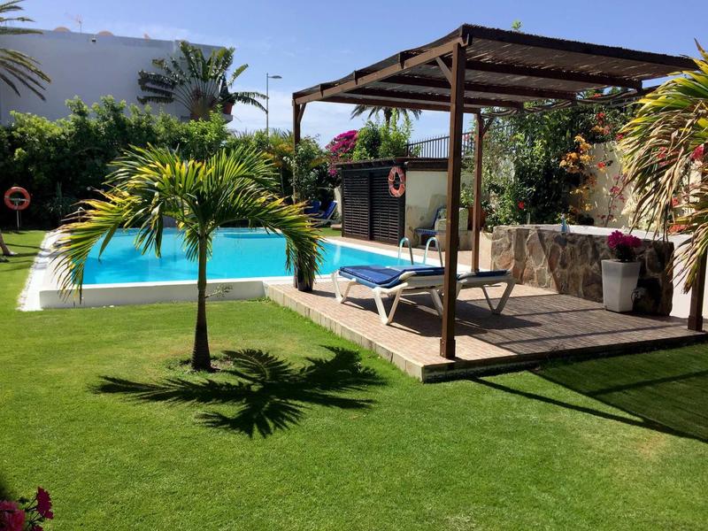 Jardin avec pelouse verte, petite piscine, pergola et chaise longue sous un ciel bleu.