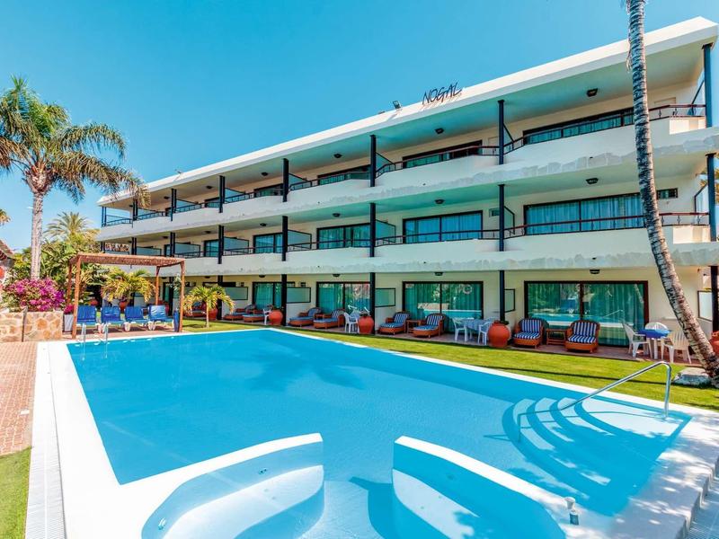 Hôtel moderne avec piscine extérieure, trois étages et palmiers sous un ciel clair.