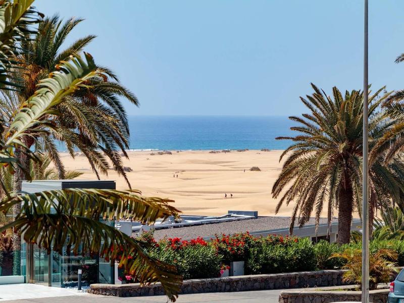 Vue depuis une terrasse avec des palmiers sur une plage de sable et la mer à l'arrière-plan.