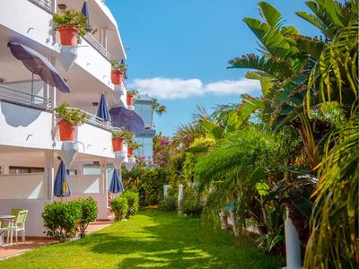 Zelená zahradní plocha vedle bílého hotelového objektu s modrými markýzami a květináči.