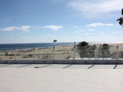 Weite Strandlandschaft mit Sanddünen, Palmen und blauem Himmel über einer weißen Terrasse.