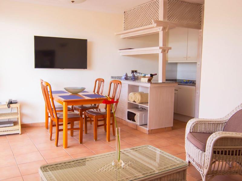 Heller Wohnraum mit Esstisch, vier Stühlen, Fernseher, Rattanmöbeln und kleiner Küchenzeile.