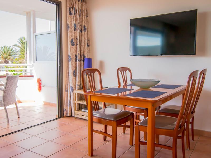 Esszimmer mit Holztisch, vier Stühlen, Wandfernseher und Blick auf Balkon mit Pflanzen.