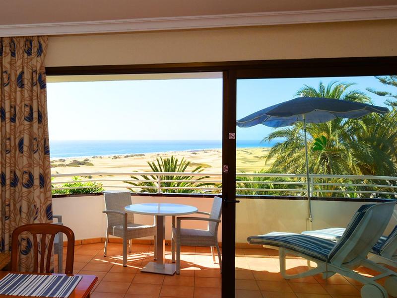 Balkon mit Tisch, Stühlen und Liegestuhl, Blick auf Meer und Palmen bei Sonnenlicht.