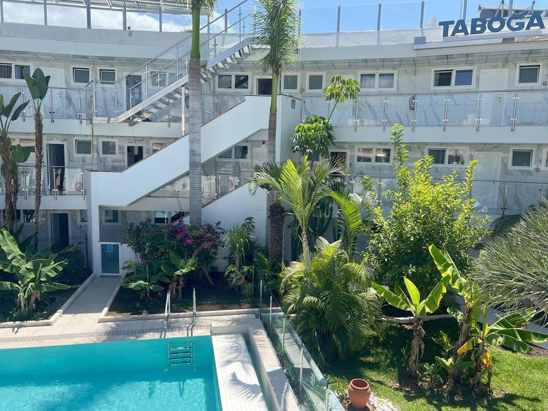 Hotelgebäude mit weißen Balkonen, Palmen und Schwimmbecken im Vordergrund.