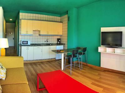 Wohnzimmer mit grünen Wänden, gelbem Sofa, rotem Tisch, Küchenzeile und TV an der Wand.