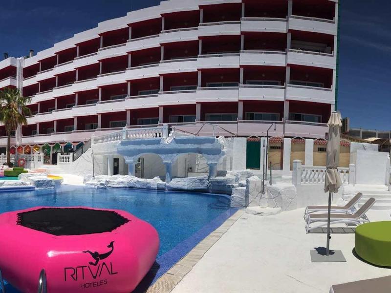 Nowoczesny basen hotelowy z różowym pływakiem, leżakami i wielopiętrowym budynkiem w tle.