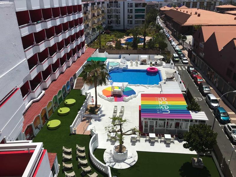 Widok na taras hotelowy z kolorowymi parasolami, leżakami i basenem otoczonym budynkami.