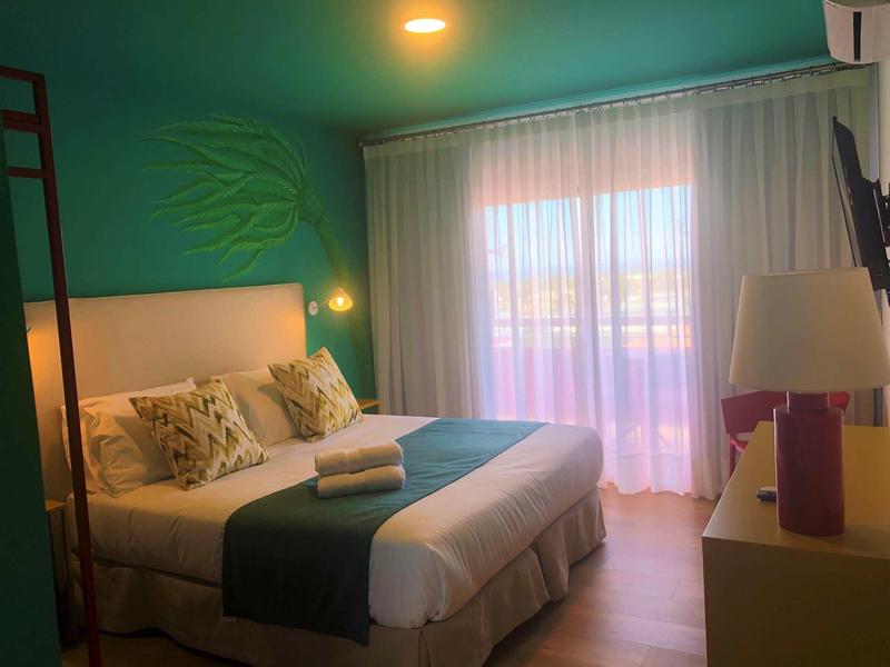 Przytulny pokój hotelowy z podwójnym łóżkiem, zieloną ścianą, balkonem i zasłonami przepuszczającymi światło.