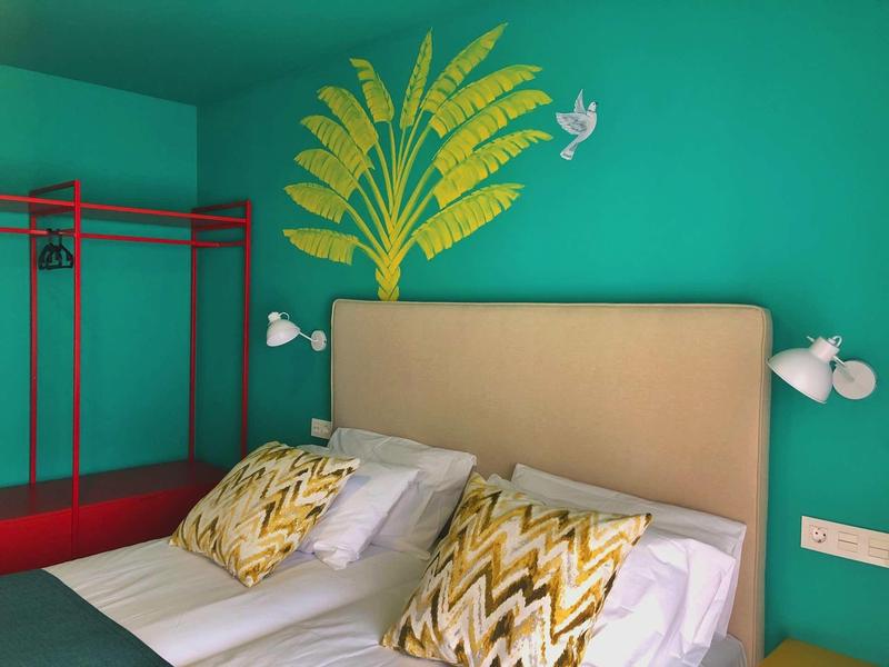 Pokój hotelowy z turkusową ścianą, beżowym zagłówkiem, wzorzystymi poduszkami i czerwonym stojakiem na ubrania.