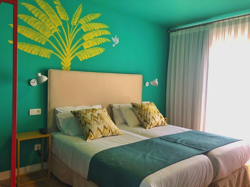 Nowoczesny pokój hotelowy z podwójnym łóżkiem, kolorowymi poduszkami i zieloną ścianą z motywem roślinnym.