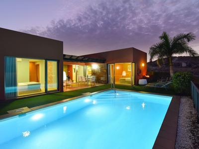 Modernes Haus mit großem beleuchtetem Pool, Glasfronten und Palmen bei Abenddämmerung.