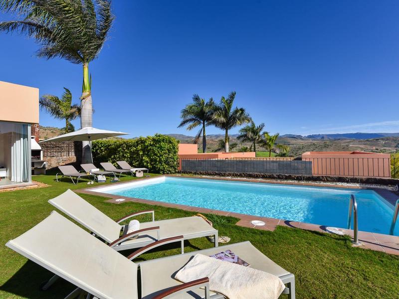 Modernes Haus mit großem Pool, Liegestühlen und Palmen unter klarem blauem Himmel.