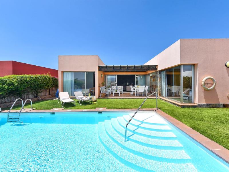 Modernes Haus mit großer Glasfront, Garten, grüner Rasenfläche und blauem Swimmingpool bei klarem Himmel.