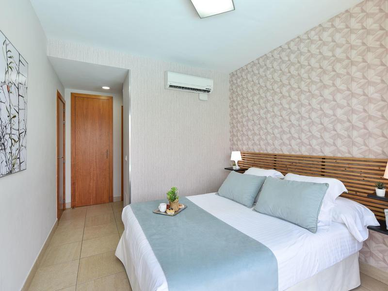 Helles Schlafzimmer mit Doppelbett, hellblauer Bettwäsche, Holzakzenten und Tapetenmuster an der Wand.