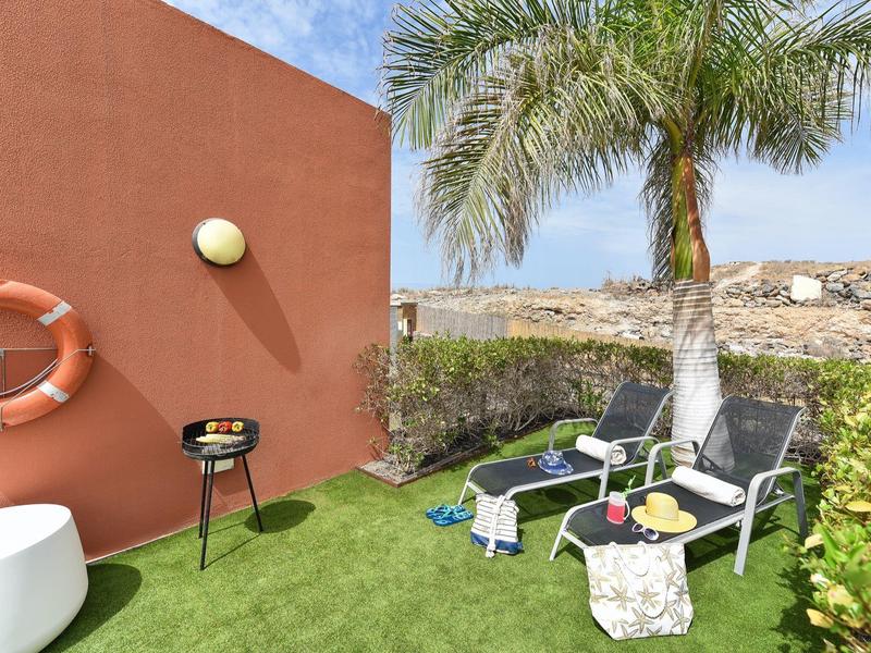 Kleiner Garten mit grüner Wiese, zwei schwarzen Liegestühlen, Palme und roter Mauer mit Rettungsring.