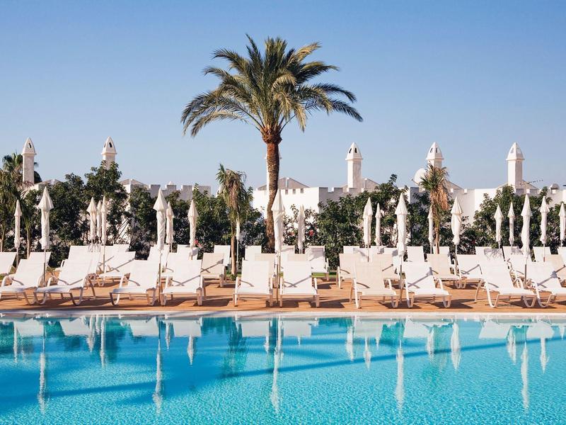 Großer Pool mit blauer Wasseroberfläche, Liegestühle und Palmen vor klarem blauem Himmel.