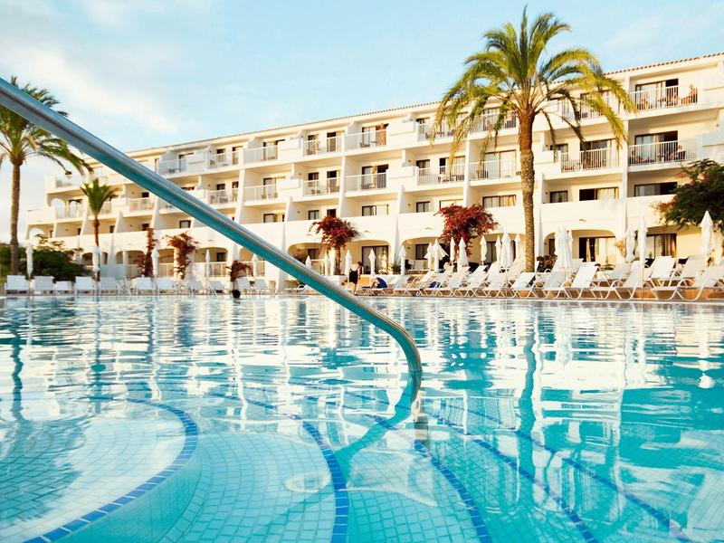 Großer blauer Hotelpool mit Metallgeländer, umgeben von Liegestühlen und Palmen bei Sonnenschein.