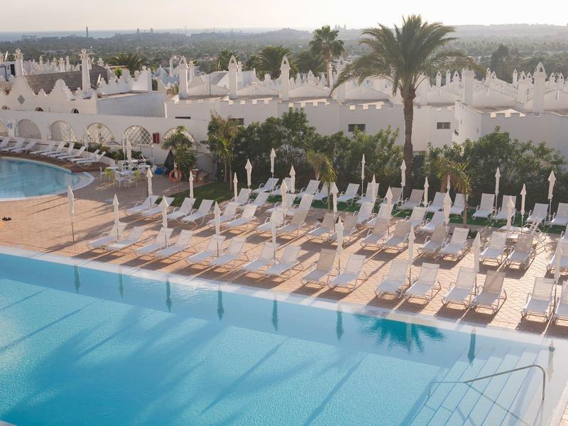 Großer Pool mit vielen Liegestühlen und weißen mediterranen Gebäuden im Hintergrund.