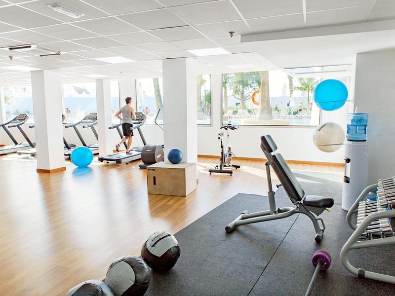 Heller Fitnessraum mit Laufbändern, Hanteln, Boxsack und Trainingsbank vor großen Fenstern.