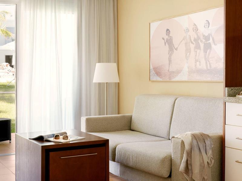 Helles Wohnzimmer mit grauem Sofa, Holztisch, Stehlampe und großem Fenster mit weißen Vorhängen.