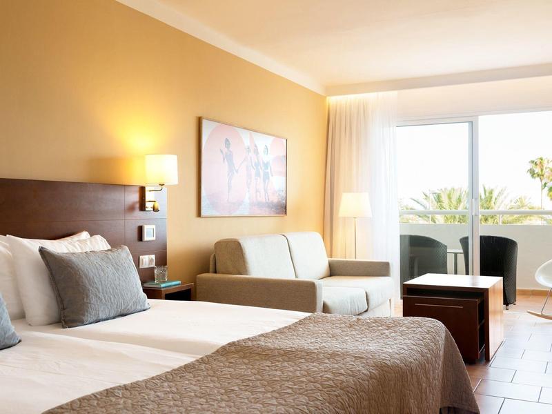 Helles Hotelzimmer mit Doppelbett, Sofa, Tisch und großem Fenster mit Blick auf Balkon und Pflanzen.