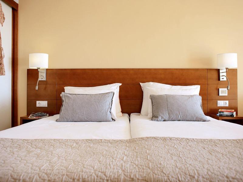 Zweibettzimmer mit braunem Holzkopfteil, weißen Kissen und beigen Decken, zwei Nachttischlampen.