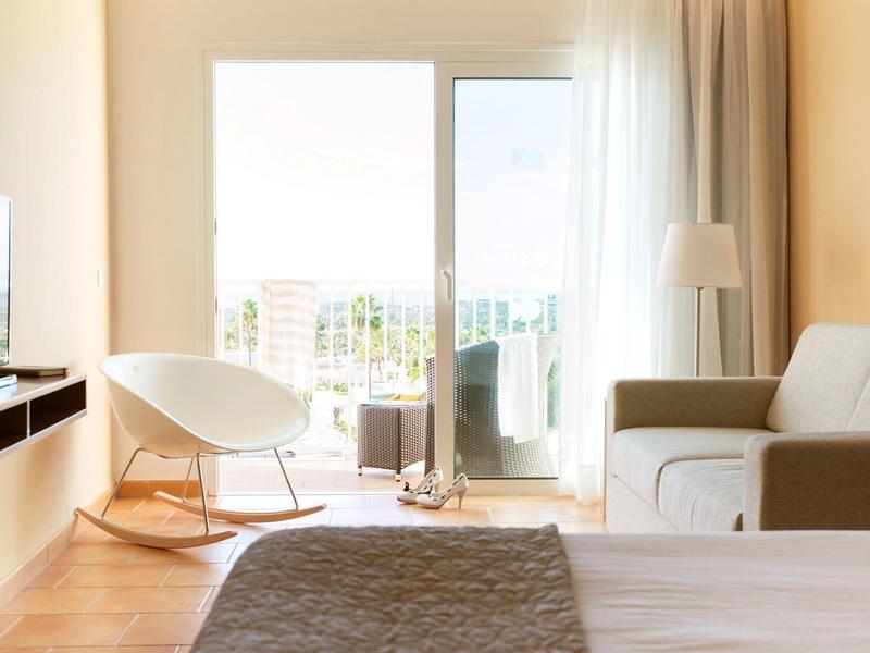 Helles Schlafzimmer mit Bett, Sessel, Sofa, großem Fenster und Blick auf Balkon und Stadt.