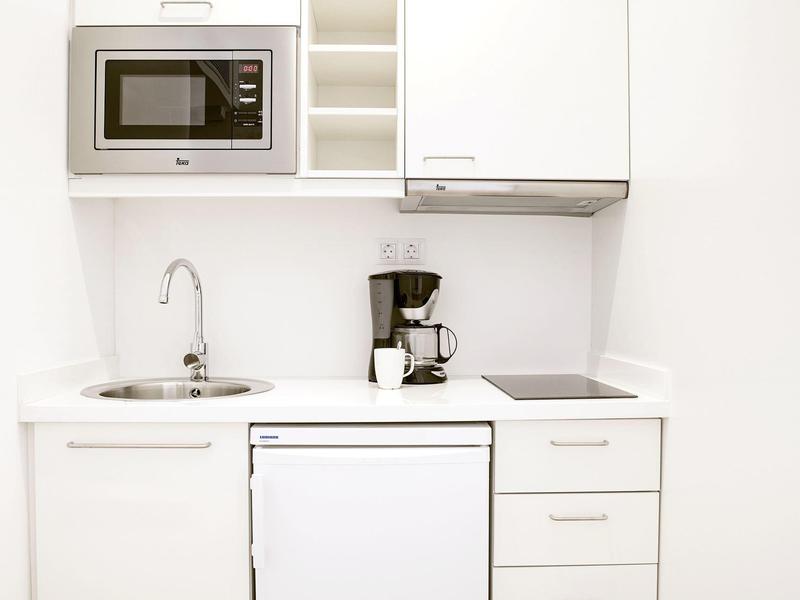 Kleine weiße Küche mit Spüle, Mikrowelle, Kaffeemaschine, Herdplatte und kleinem Kühlschrank.