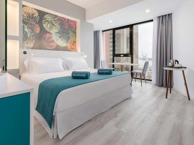 Chambre d'hôtel moderne avec lit double, draps blancs, déco turquoise et vue sur balcon.