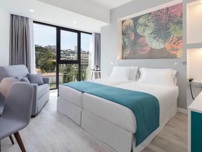 Chambre d'hôtel lumineuse avec lit double, fauteuils et balcon donnant sur la nature.