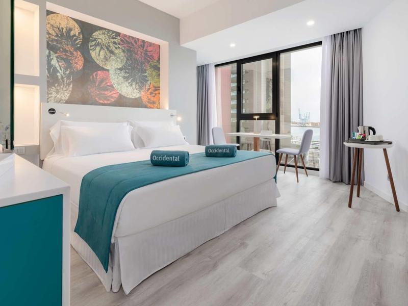 Chambre d'hôtel moderne avec lit double, draps blancs, déco turquoise et vue sur balcon.