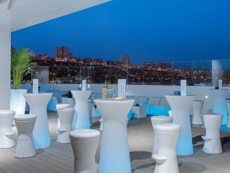 Bar extérieur avec tables et tabourets blancs offrant une vue nocturne sur la ville.