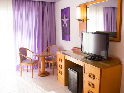 Jasny pokój hotelowy z fioletowymi zasłonami, dwoma krzesłami i telewizorem na komodzie.