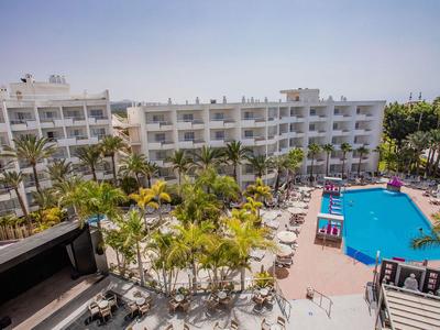 Hotel complex with large outdoor pool, sun loungers, and palm trees under clear sky.