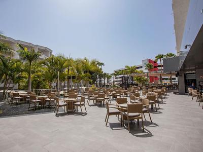 Duży taras na zewnątrz z wieloma stołami i krzesłami, otoczony palmami pod czystym niebem.