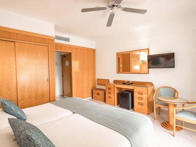 Jasny pokój hotelowy z dwoma łóżkami, biurkiem, krzesłami i telewizorem na ścianie.