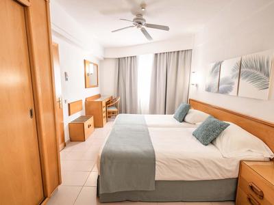 Jasny pokój hotelowy z łóżkiem podwójnym, wentylatorem sufitowym i zasłonami przy dużym oknie.