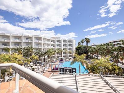 Hotel z dużym basenem otoczonym palmami i białymi budynkami pod niebieskim niebem.