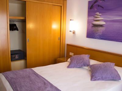 Pokój hotelowy z łóżkiem podwójnym, fioletowymi poduszkami i narzutą, otwartą szafą i relaksującą dekoracją ścienną.